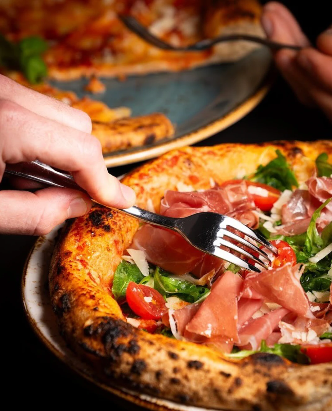 Nahaufnahme: Gabel nimmt Stück Pizza mit Prosciutto, Rucola & Tomaten.